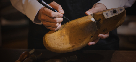 オーダーメイドシューズの製作風景。熟練の靴職人が木型（ラスト）を手に持ち、顧客のためのデザインラインをペンで精密に描き込んでいる。伝統的な手仕事とクラフトマンシップを象徴する一場面。