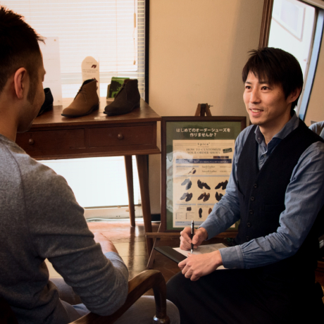店内で、男性店員が顧客と靴について相談している様子。店員は笑顔で筆記用具を持ち、顧客は店員の方を向いている。奥には靴がディスプレイされたテーブルが見える。
