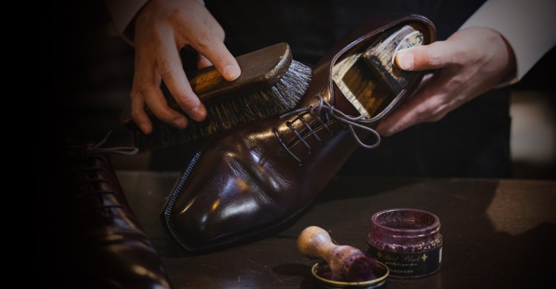 暗く落ち着いた雰囲気の中、職人が光沢のあるバーガンディ色の革靴を手に持ち、ブラシで丁寧に磨き上げている様子。手元には靴クリームの瓶も見える。