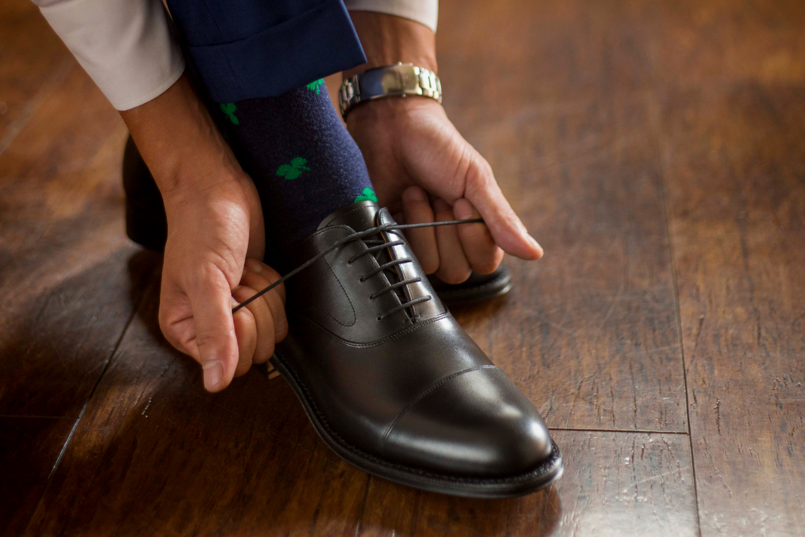 スーツを着た男性が、光沢のある木製の床の上で、黒い革靴の靴紐を結んでいる手元と足元のクローズアップ。紺色の靴下には緑色の模様が入っている。