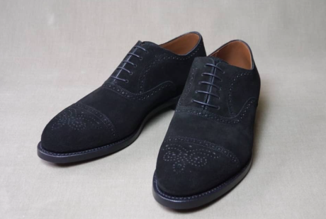 グレーの無地の背景に置かれた、黒いスエード製のセミブローグシューズ一足。つま先のキャップ部分にはメダリオン、縫い目にはパーフォレーションという穴飾りが施されている。