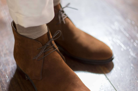 白いパンツにベージュの靴下を合わせ、ブラウンのスエード製チャッカブーツを履いた人物の足元。光沢のある床に靴が映り込んでいる。
