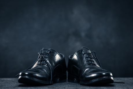 ローアングルから捉えた、少し履き古された一対の黒い革製ドレスシューズ。ドラマチックな暗い背景の前に置かれている。