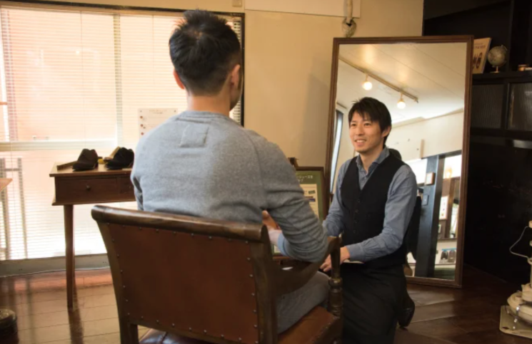 日当たりの良いオーダーメイドシューズの店舗内で、革張りの重厚な椅子に座る顧客（後ろ姿）に対し、ベストを着用した店員が、顧客と目線を合わせるように屈みながら、温かい笑顔で丁寧に接客・カウンセリングを行っている。大きな鏡には、親身に対応する店員の親しみやすい表情がはっきりと映し出されている。