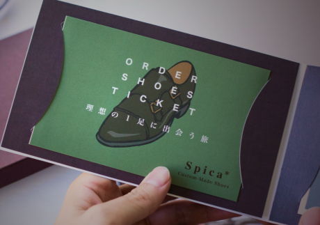 手に持たれた「ORDER SHOES TICKET」と書かれた緑色の冊子。イラストの靴と「理想の1足に出会う旅」という日本語、そしてブランド名「Spica Custom-Made Shoes」がデザインされている。