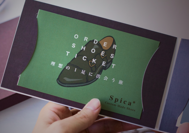 手に持たれた「ORDER SHOES TICKET」と書かれた緑色の冊子。イラストの靴と「理想の1足に出会う旅」という日本語、そしてブランド名「Spica Custom-Made Shoes」がデザインされている。