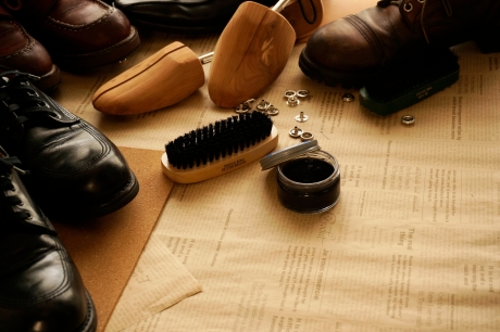 新聞紙の上に、黒い革靴、シューツリー、靴ブラシ、靴墨、靴紐の金具などの靴手入れ用品が配置されている様子