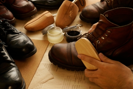 茶色のレザーブーツを手入れしている人の手元。木製のブラシでブーツを磨き、周りにはシューツリーが入った他の革靴や、様々なシューケア用品(クリーム、ワックスなど)が置かれている。