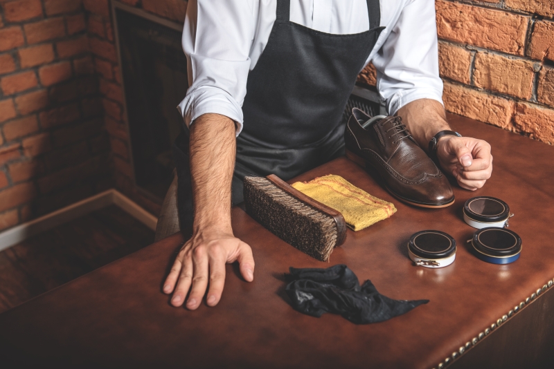 エプロンを着た靴磨き職人が木製のテーブルで茶色の革靴を手入れしている様子。テーブルには大きなブラシ、布、複数のシュークリーム缶が並べられている。