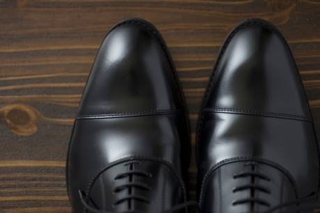 艶やかに磨き上げられた黒色の革靴(ビジネスシューズ)のつま先部分。木目調の背景に置かれ、美しい光沢を放っている。