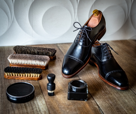 木製の表面に、黒い革の紳士靴(シューツリー入り)が2足、シューブラシが3本、靴磨き用のクリームとブラシが置かれている。背景は白と灰色の幾何学模様の壁。これは靴の手入れやメンテナンス用品を構成するイメージ。