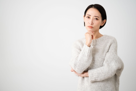 白い背景を背に、灰色のニットを着た短髪の女性が、顎に手を当てて考え込んでいる様子。彼女はカメラを見つめ、思慮深い表情をしている。