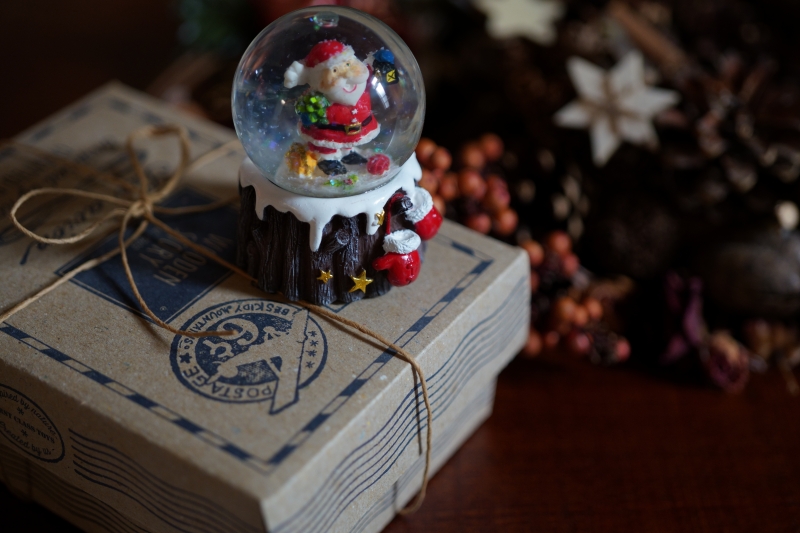 ラッピングされた小包の上にサンタクロースのフィギュアが入ったスノードームが置かれている。背景にはクリスマスらしい飾り付けが施されている。