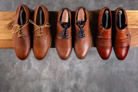 木製の棚に整然と並べられた6足のメンズ革製ドレスシューズの俯瞰図。それぞれが異なるブラウンの色合いとスタイルを持ち、シューツリーが挿入されている。左から順に、ライトブラウンのブローグダービー、ミディアムブラウンの青い靴紐ダービー、そしてダークブラウンのダブルモンクストラップシューズが写っており、背景はコンクリート調のグレーの床である。