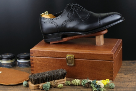 黒い背景の前に置かれた木製の靴磨き箱の上に、木製スタンドで高くディスプレイされた黒い革製のキャップトゥブローグドレスシューズ。シューズには金色のシューツリーが挿入されている。手前左側には、靴磨き缶と茶色の磨き布があり、中央には靴磨きブラシと装飾用の松ぼっくりや小枝が散らばっている。
