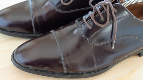 明るい木製の表面に置かれた、埃や白い汚れが付着し手入れが必要な状態のダークブラウンの革製ドレスシューズのクローズアップ。右側の靴にはシューツリーが装着されている。