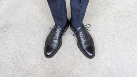 黒い革のストレートチップ（内羽根式）紳士靴とダークスーツのズボンを着用した足元が、コンクリートの床に立っている様子。