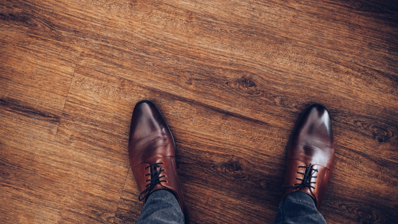 木製の床に立つ男性の足元を上から見た写真で、茶色の革製ドレスシューズとダークなパンツが写っています。