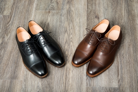 木製の床に置かれた、黒と茶色の高級紳士用革製ドレスシューズ（キャップトゥ）が2足ずつ並べられています。ビジネスやフォーマルなシーンに最適な、クラシックなデザインのメンズシューズです。