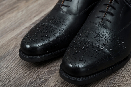 木目の床に置かれた黒い革製ドレスシューズのつま先部分のクローズアップ。靴の撥水性を示すように、表面には多数の水滴が付着している。